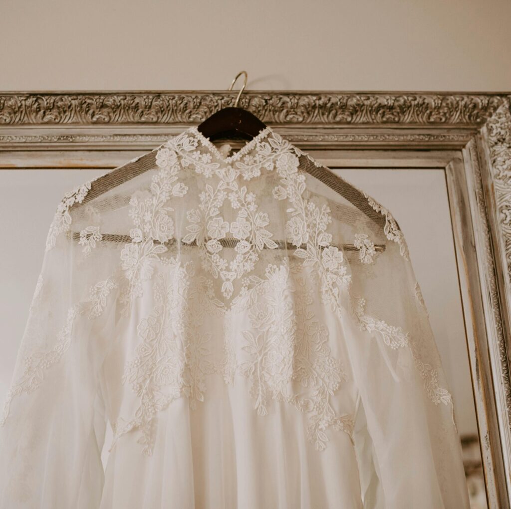 A delicate lace wedding gown hanging on a decorative mirror indoors.